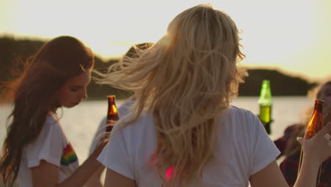 A-blonde-female-in-white-t-shirt-is-moving-her-slender-body-and-hands-on-the-beach-party-on-the-sand-river-coast-with-beer.